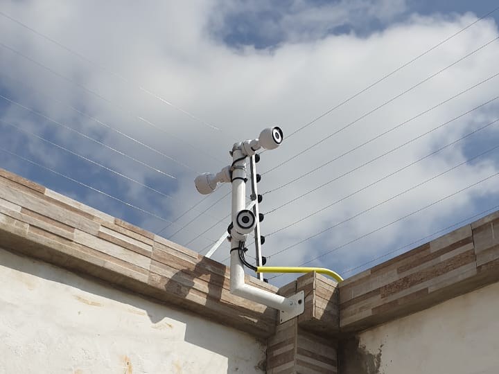 Two bullet cameras and a dome on a single bracket at a boundary wall corner, below an electric fence line
